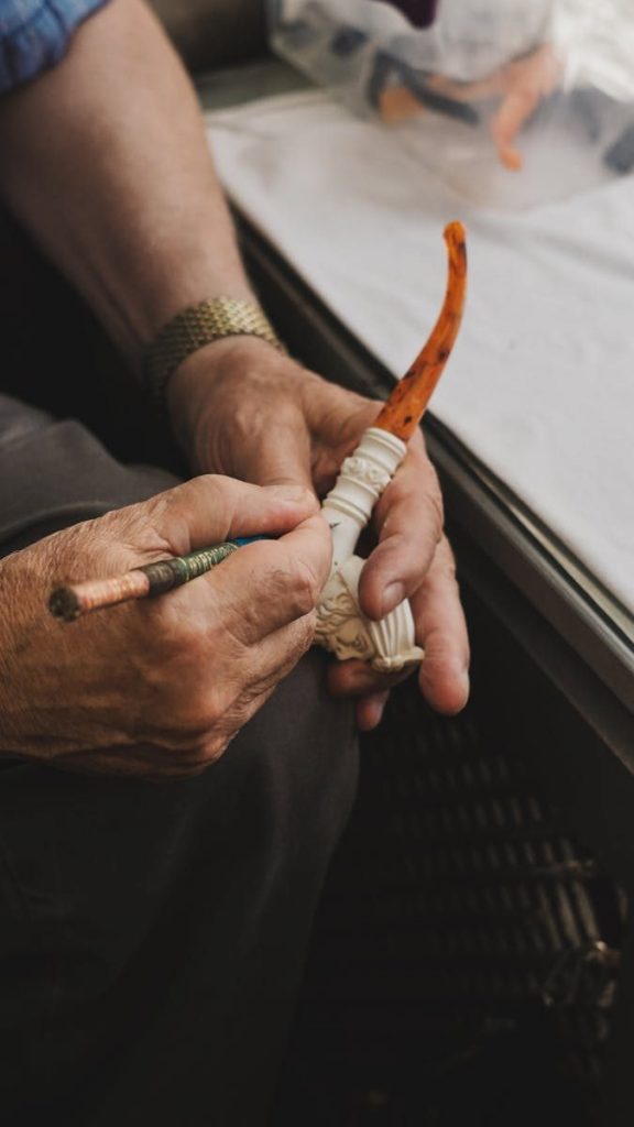An artisan meticulously carves and paints a handmade meerschaum pipe indoors, showcasing skill and craftsmanship.